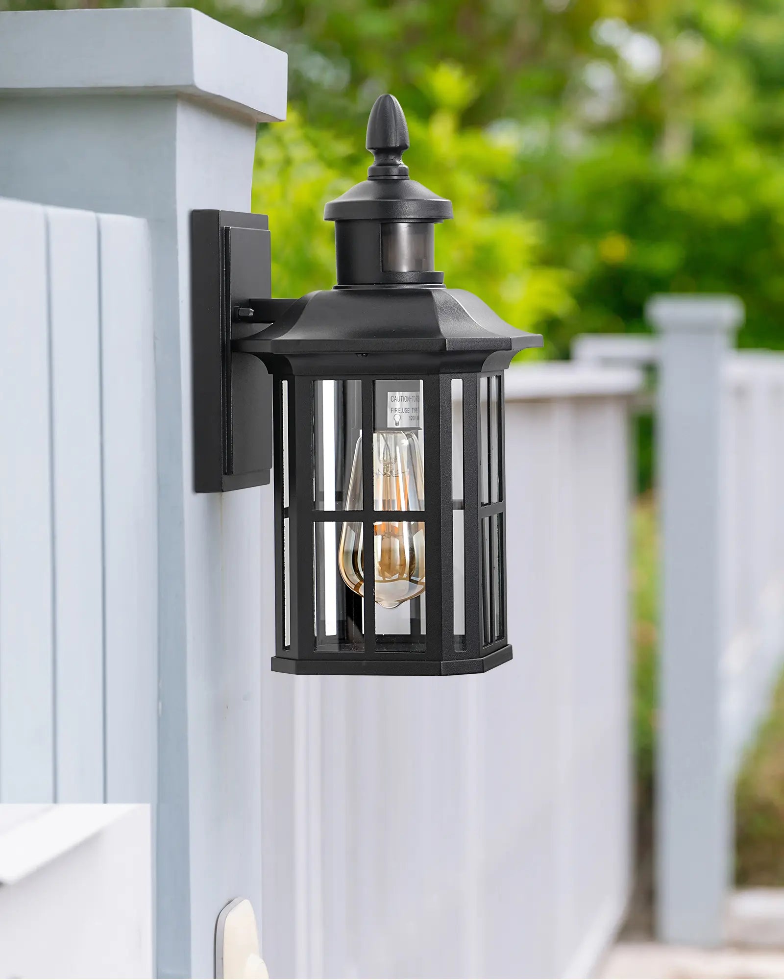 Motion Sensor Dusk to Dawn Outdoor Wall Lights  on a white wooden post with greenery in the background