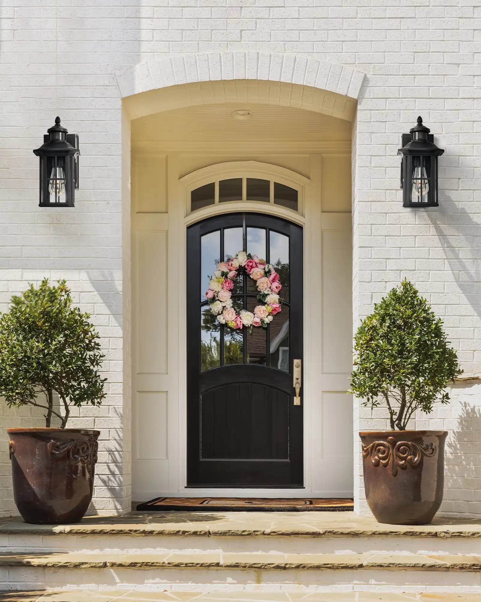 Front door with floral wreath, flanked by two potted plants on a white brick wall.