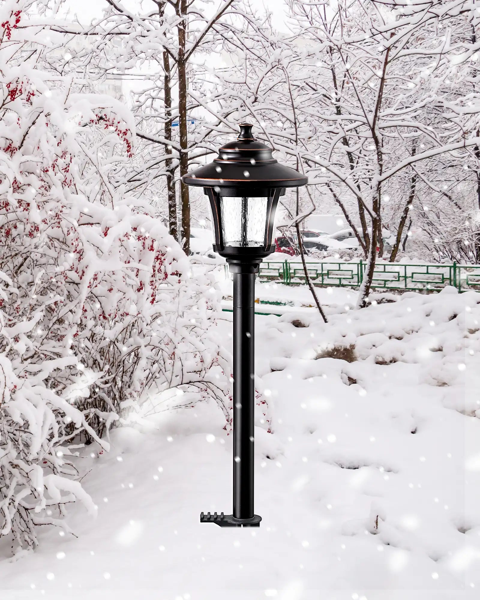 Black outdoor lamp post in a snowy landscape with trees and a bench in the background.