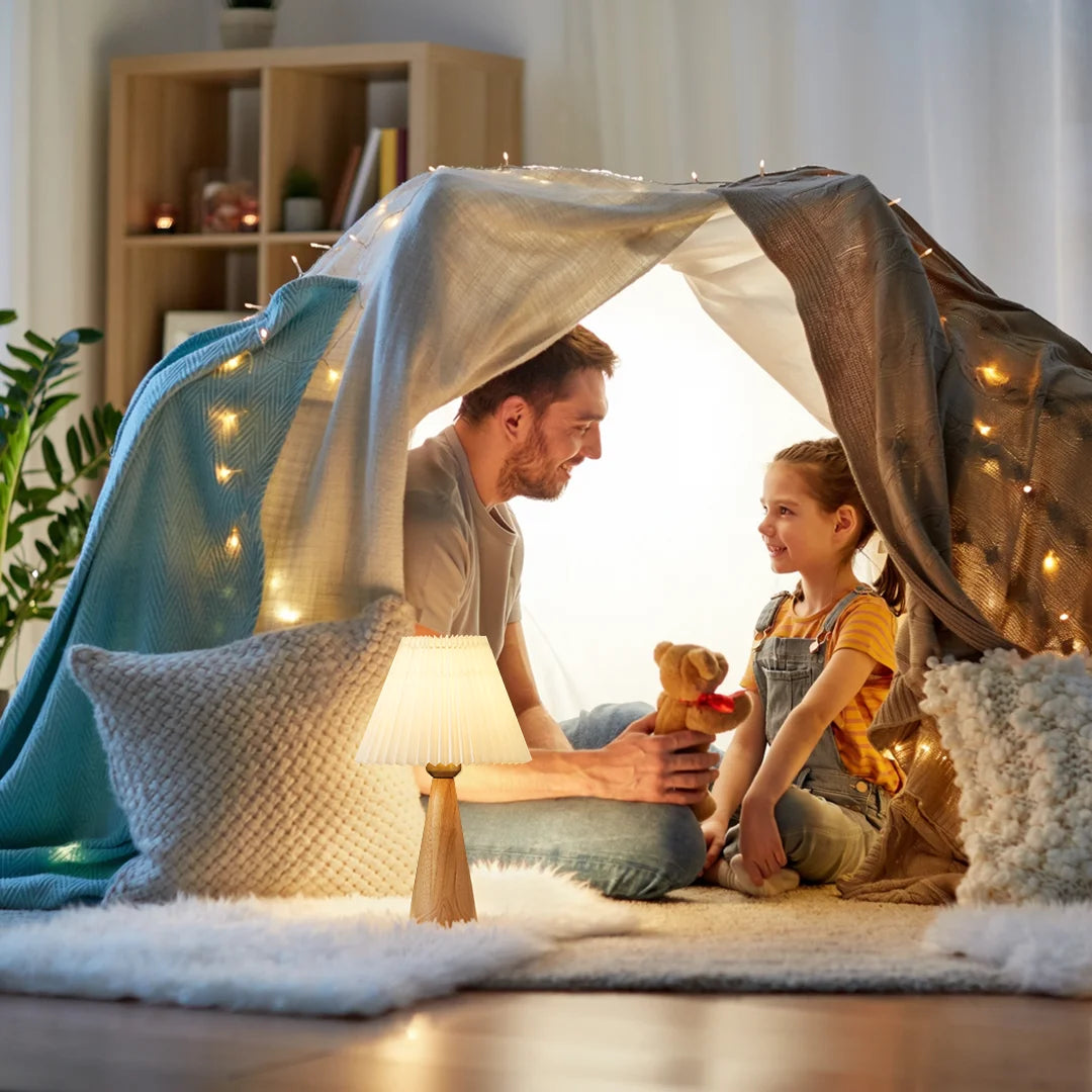 Man and child playing under a homemade fort with string lights in a cozy room.