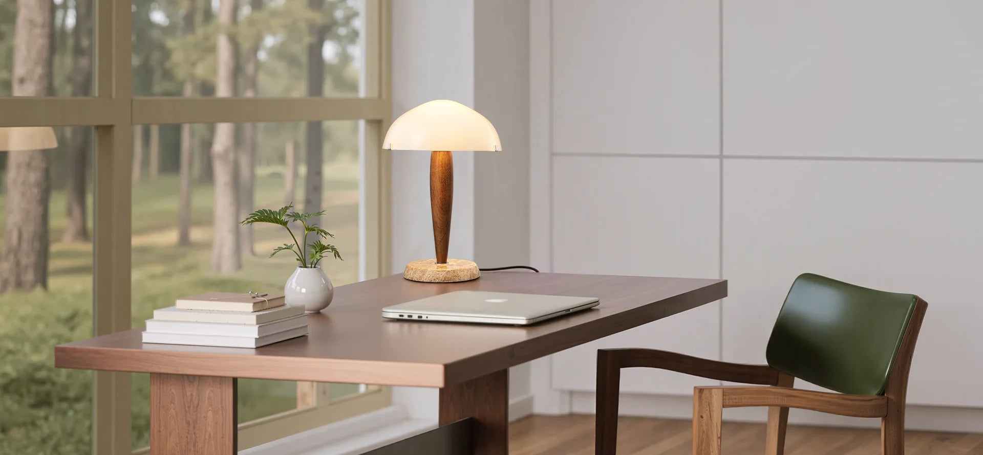 Modern desk with lamp, books, and plant in a room with large windows showing greenery outside.