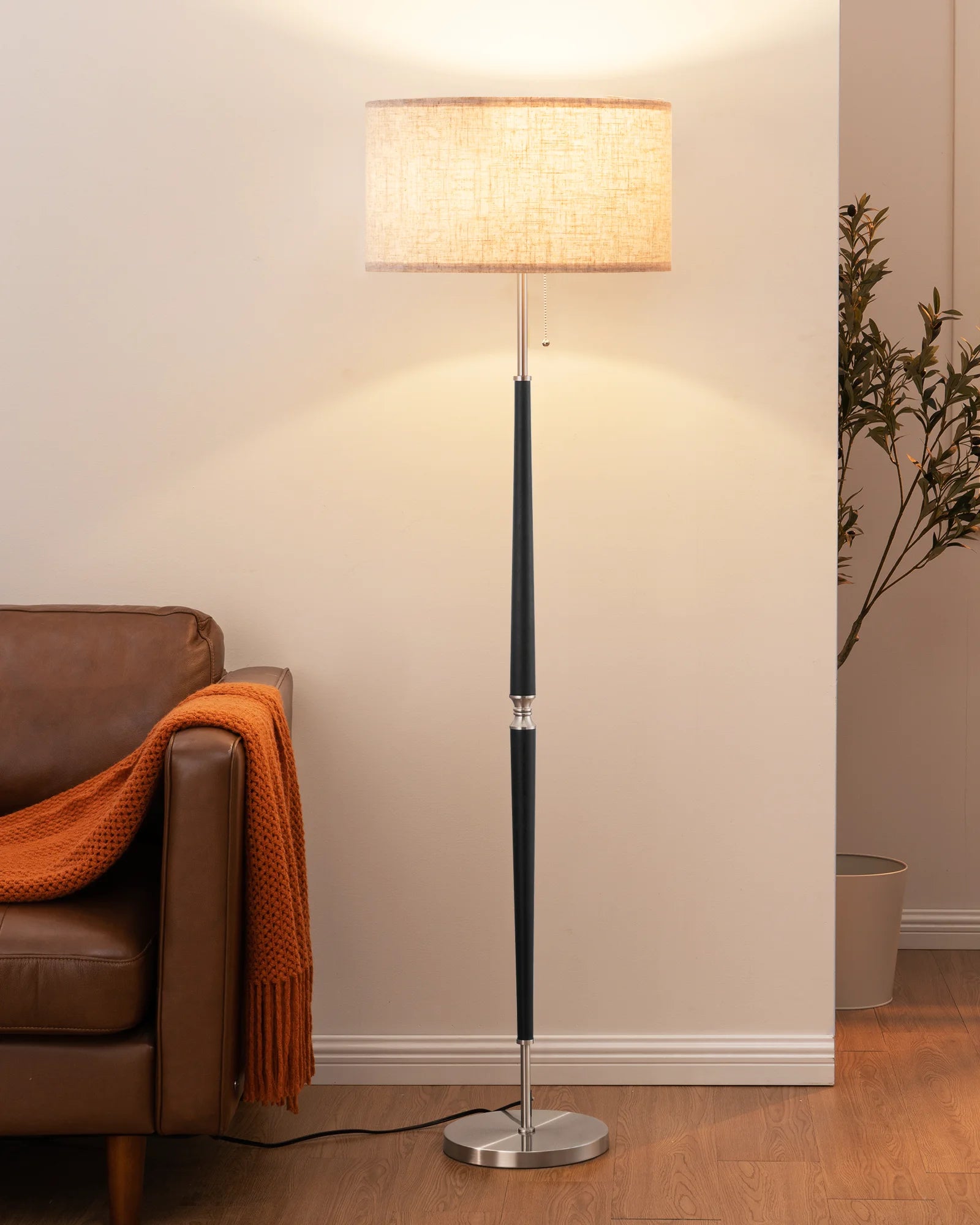 Floor lamp with a beige shade next to a brown leather sofa in a living room setting.