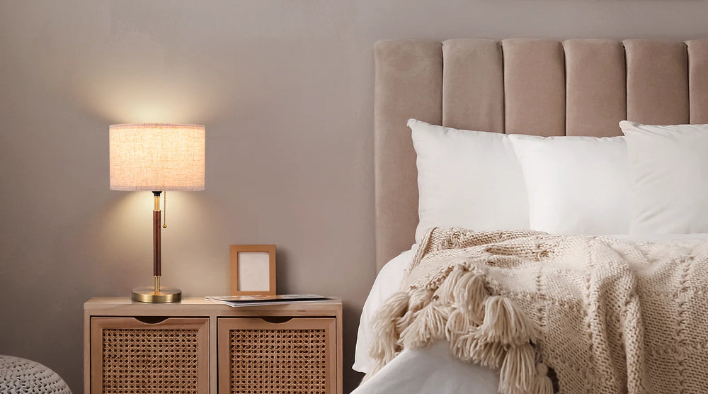 Neatly made bed with beige headboard and blanket, nightstand with lamp and decor items.