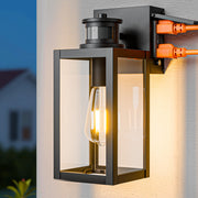 A modern outdoor wall sconce with a black metal frame, clear glass panels, and a glowing warm Edison bulb. It is mounted on a white stucco wall with a blurred background showing a house at dusk and some greenery.
