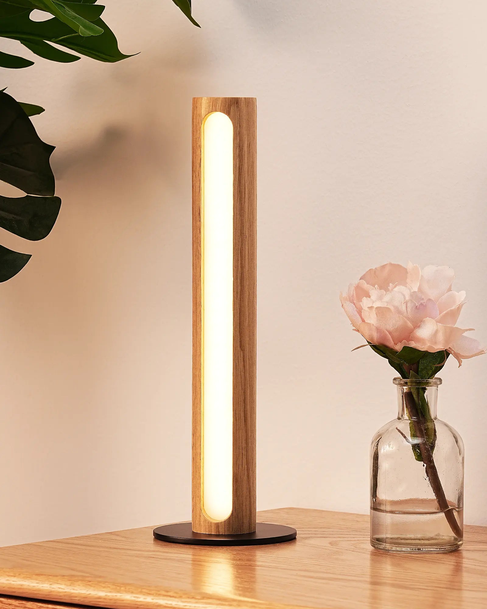 Minimalist Wood Cylinder Table Lamp on a wooden surface with a vase of flowers and leaves in the background