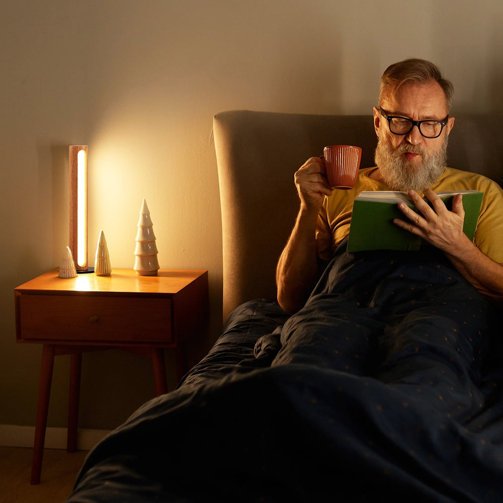 Man reading a book and holding a mug in a cozy room with warm Minimalist Wood Table lighting.
