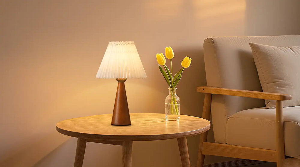 Round wooden table with a lamp and vase of yellow tulips next to a beige armchair.