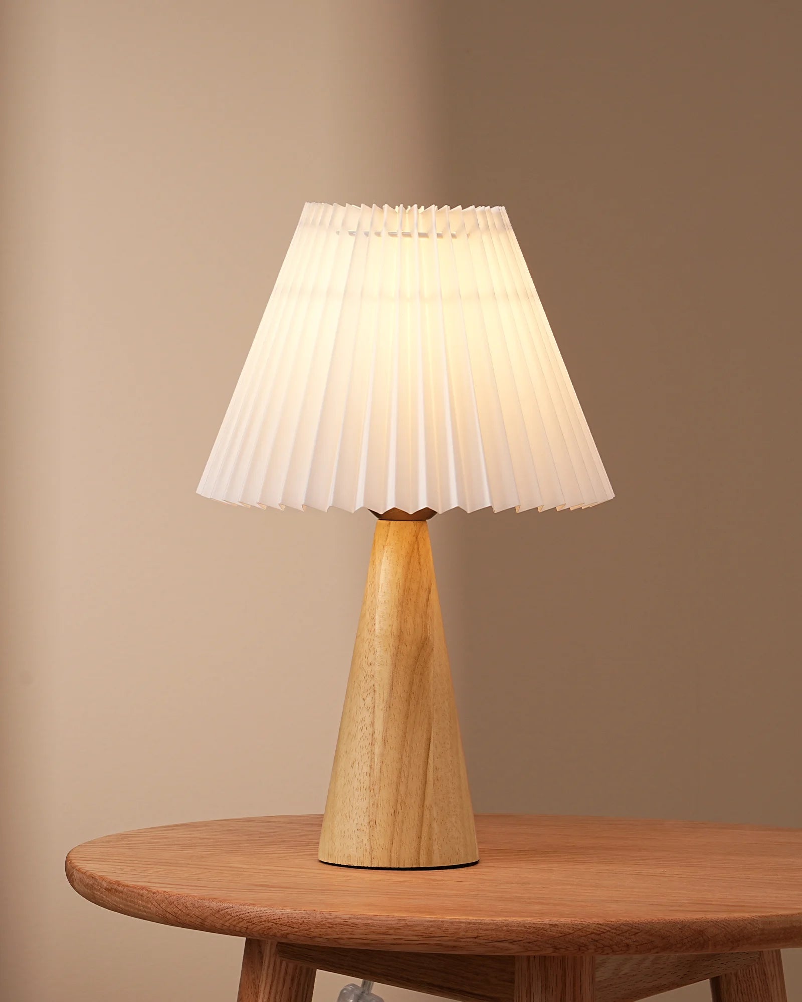 Sustainable Natural  Pleated White Table Lamp with pleated white shade on a wooden base, placed on a wooden table against a beige wall.