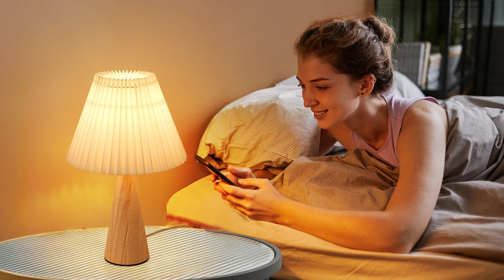 Woman lying in bed using a smartphone with a Pleated White Table Lamp beside her