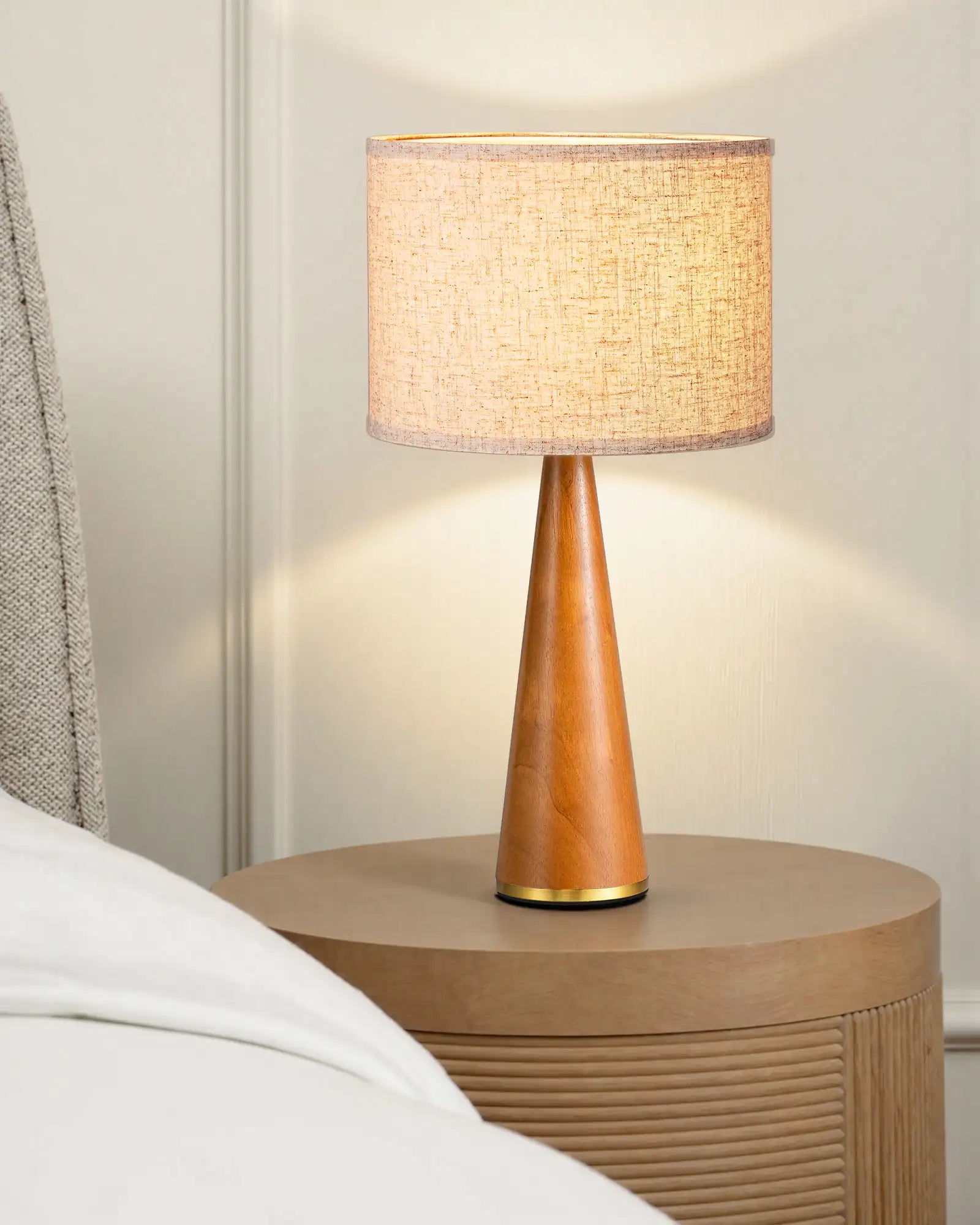 Table lamp with wooden base and beige shade on a nightstand next to a bed.