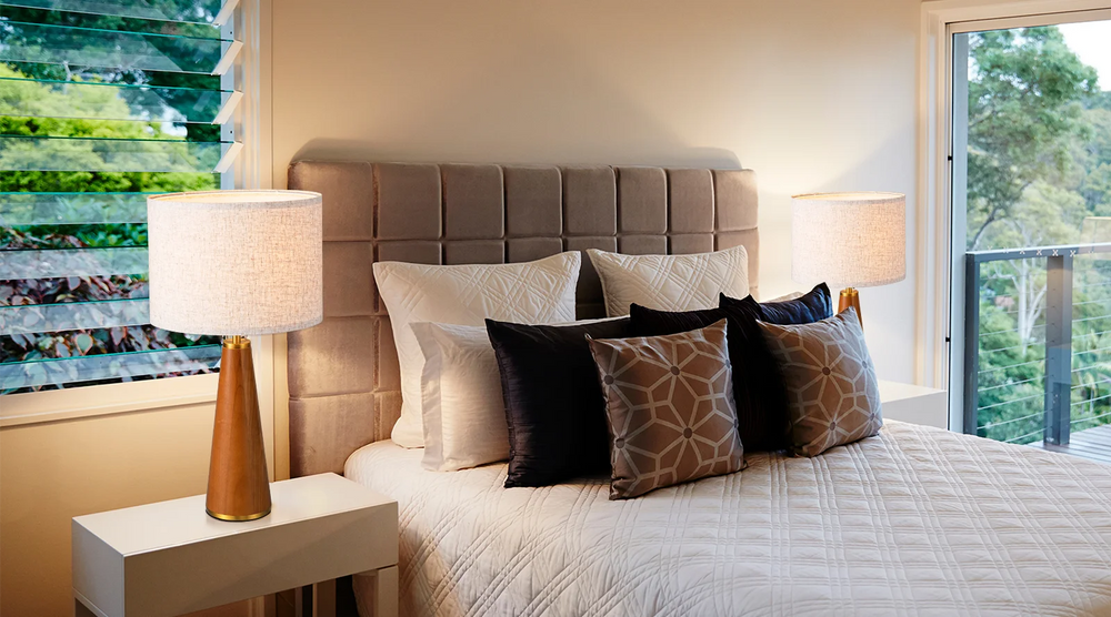 Bedroom with a bed featuring patterned pillows, nightstands with a pair of Solid Wood Table Lamps, and large windows.