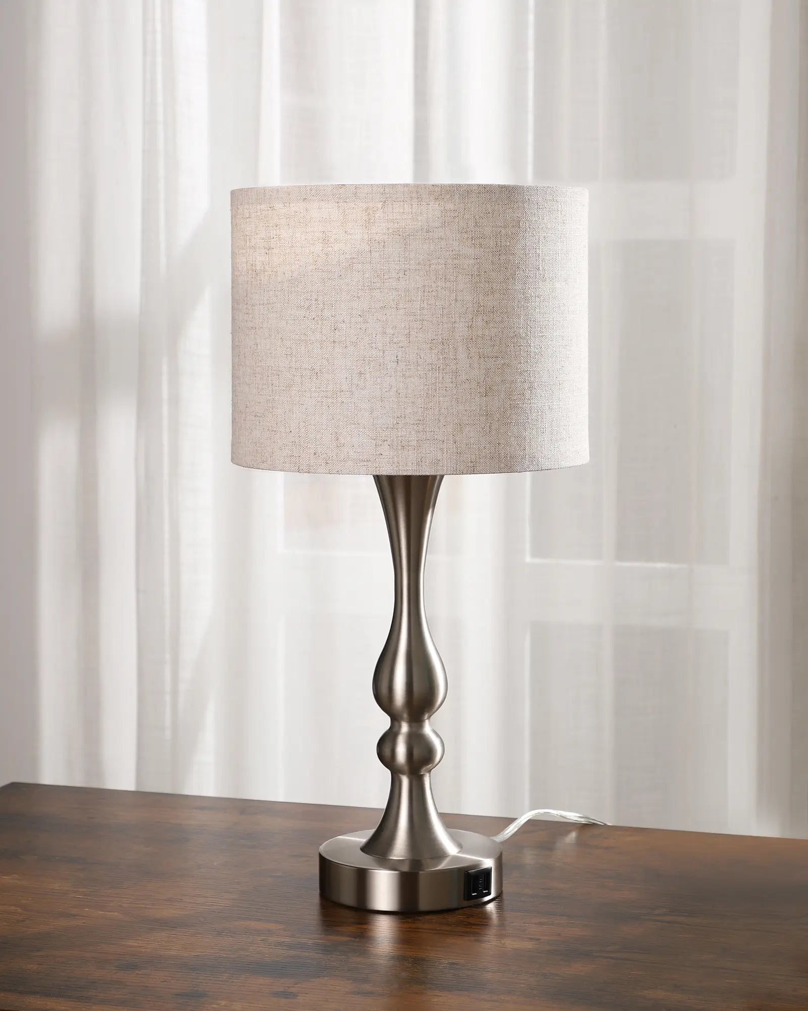 Silver Traditional Table Lamp with a beige shade on a wooden surface in front of a white curtain